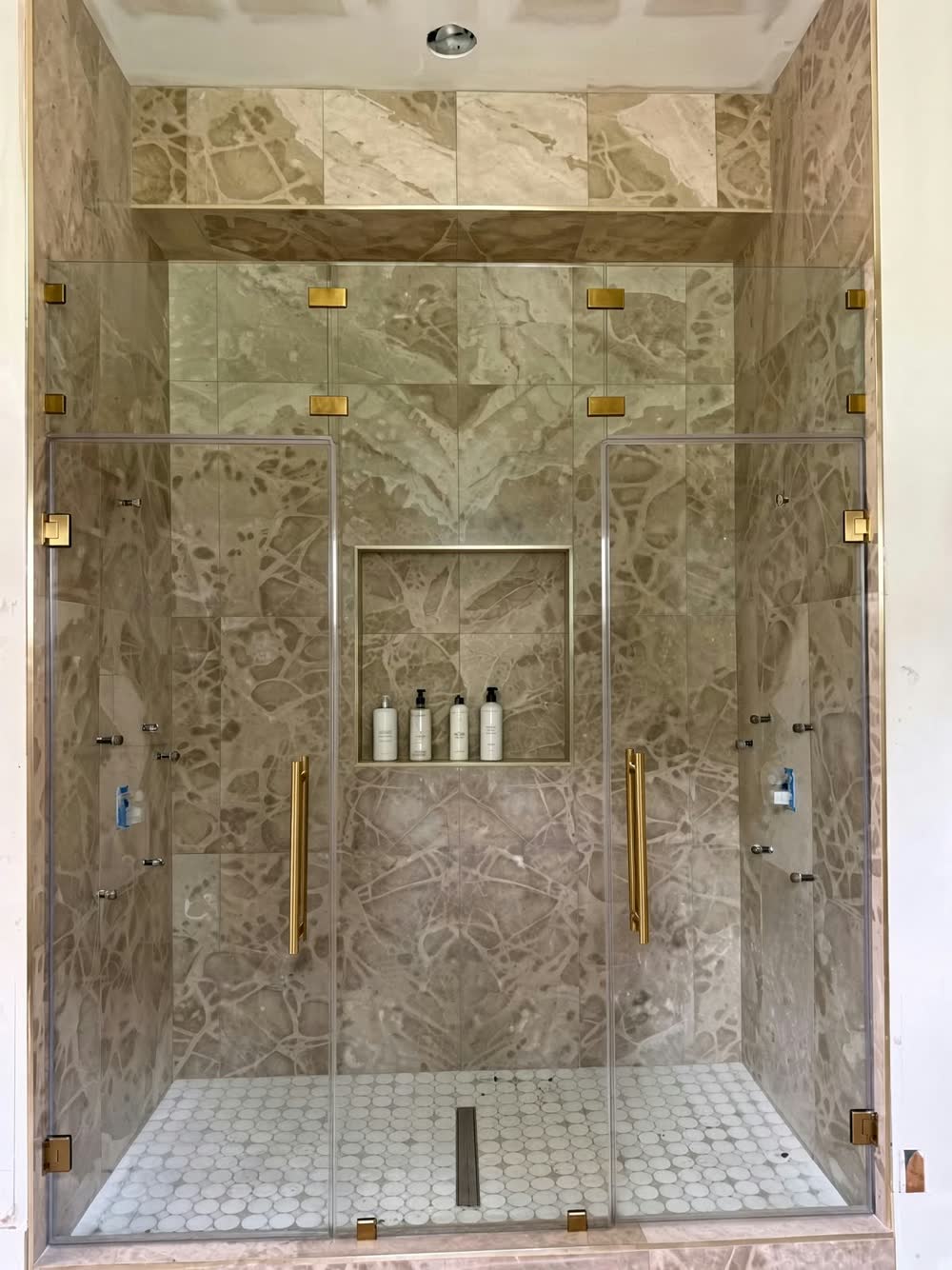 Large walk-in shower with double frameless glass doors, stone-look tile, a lit niche and brass hardware