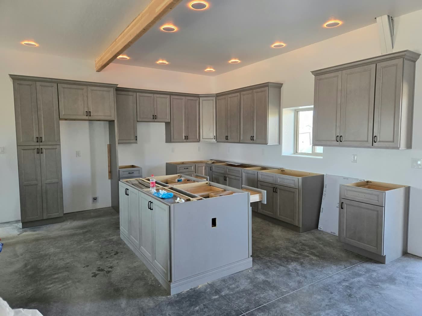 Gray kitchen cabinets and island being installed under recessed lights and a wood ceiling beam, before countertops go on