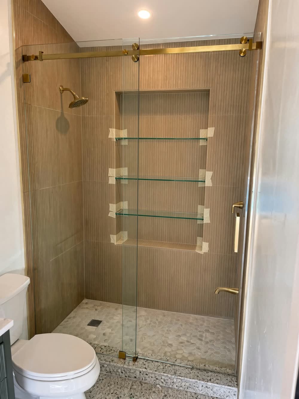 Sliding glass shower door with brass hardware, wood-look wall tile, glass shelves and a pebble floor — ProRock Remodeling, Bay Area